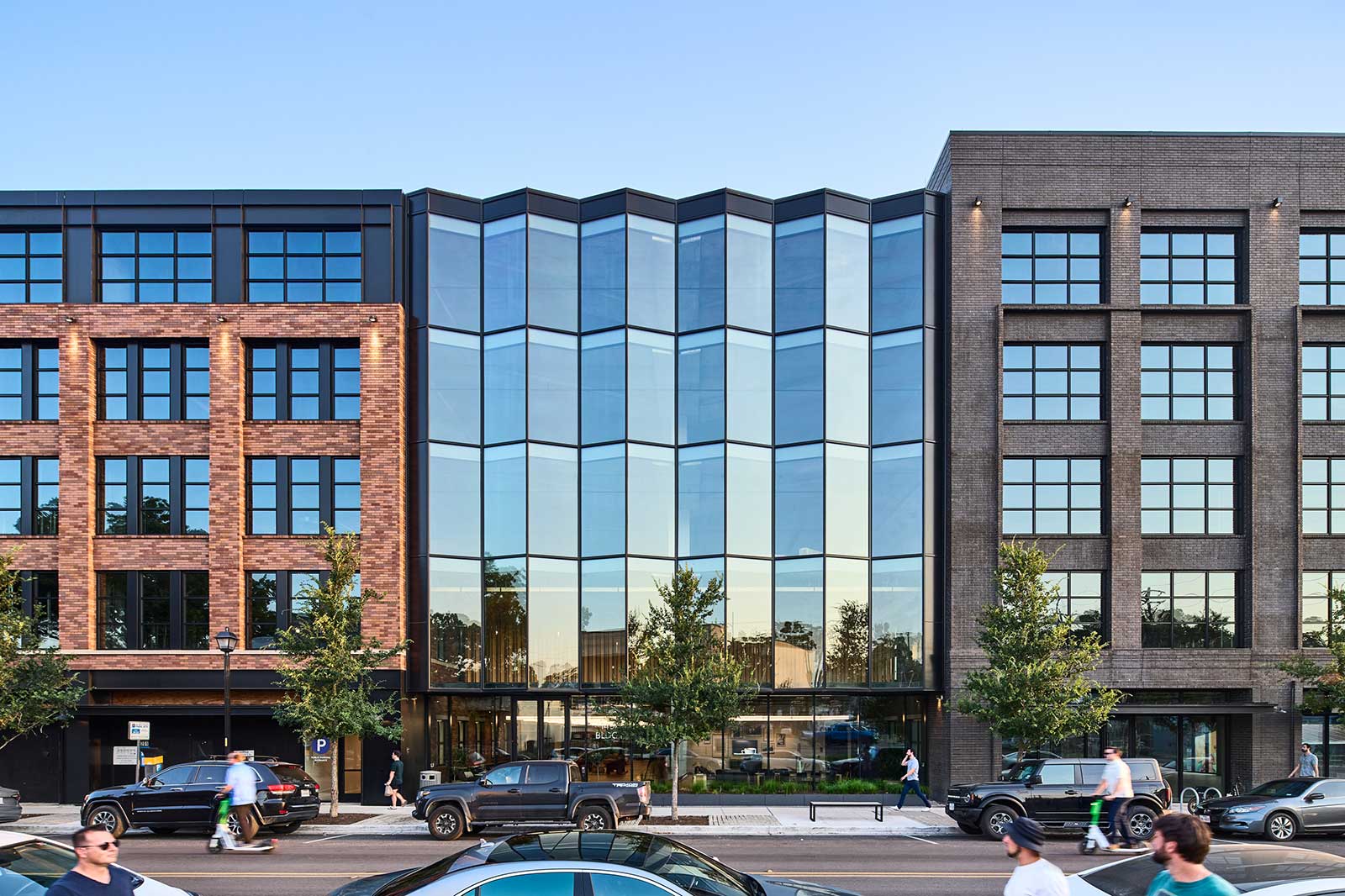 A modern building featuring a mix of glass and brick with large windows and greenery in front, alongside a busy street.