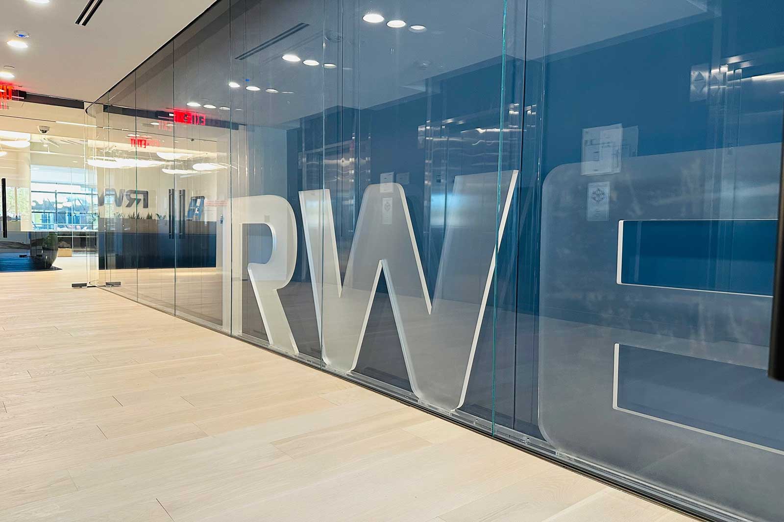 An office interior featuring large transparent letters 'RWE' on glass walls, modern design, and wooden flooring.